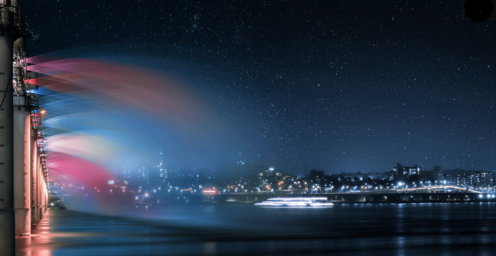 Travel to tour the colored fountain show at the Banpo Bridge Rainbow Fountain against the water reflecting a starlit cityscape of Seoul, South Korea.