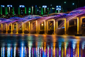 Travel to tour the colored fountain show of the Banpo Bridge Rainbow Fountain against the water reflecting the starlit cityscape of Seoul, South Korea.