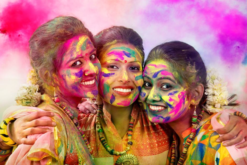 a group of 3 indian women embrace while colorful powder is tossed all around them to celebrate one of the best and most must see festivals in the world, Holi.