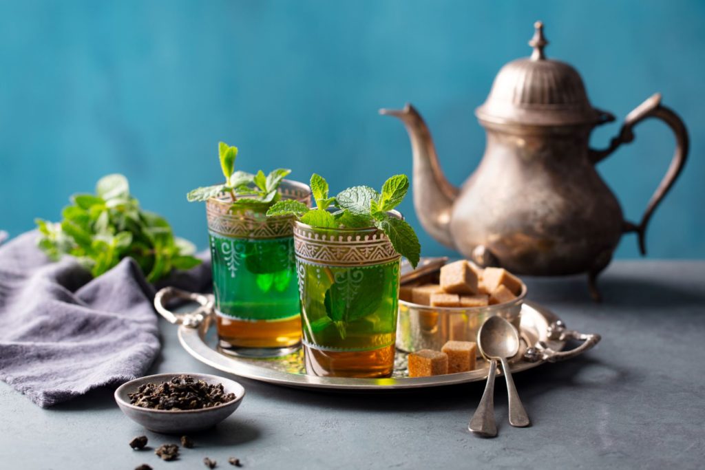 Two cups of Moroccan mint tea garnished sit on a tray with sugar cubes next to a tea pot in the background. Visit Morocco to drink traditional tea with Jaya Travel & Tours.