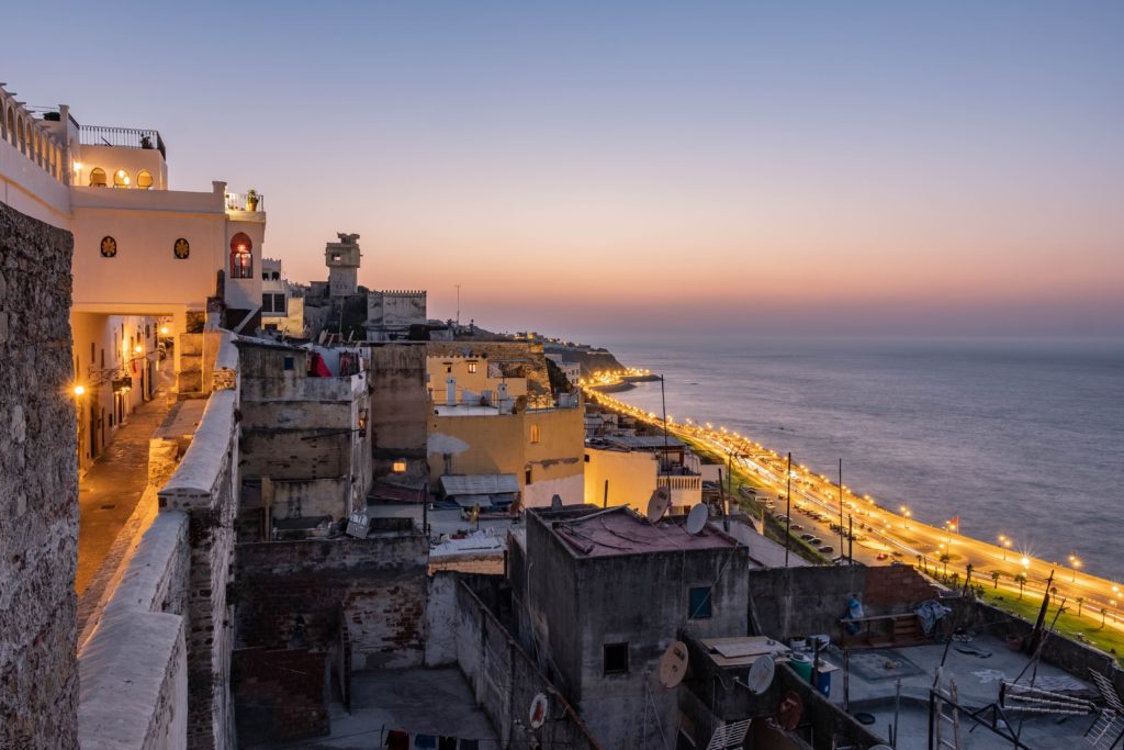 Water washes onto the sand on one side and a beautiful coastal city sits on the other at dusk in Tangier, Morocco, which you can see on a movie tour with Jaya Travel & Tours.