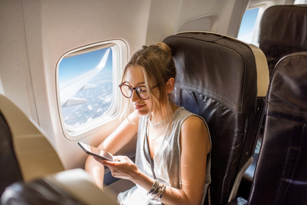 a woman sits on an airplane in a window seat while on her phone.