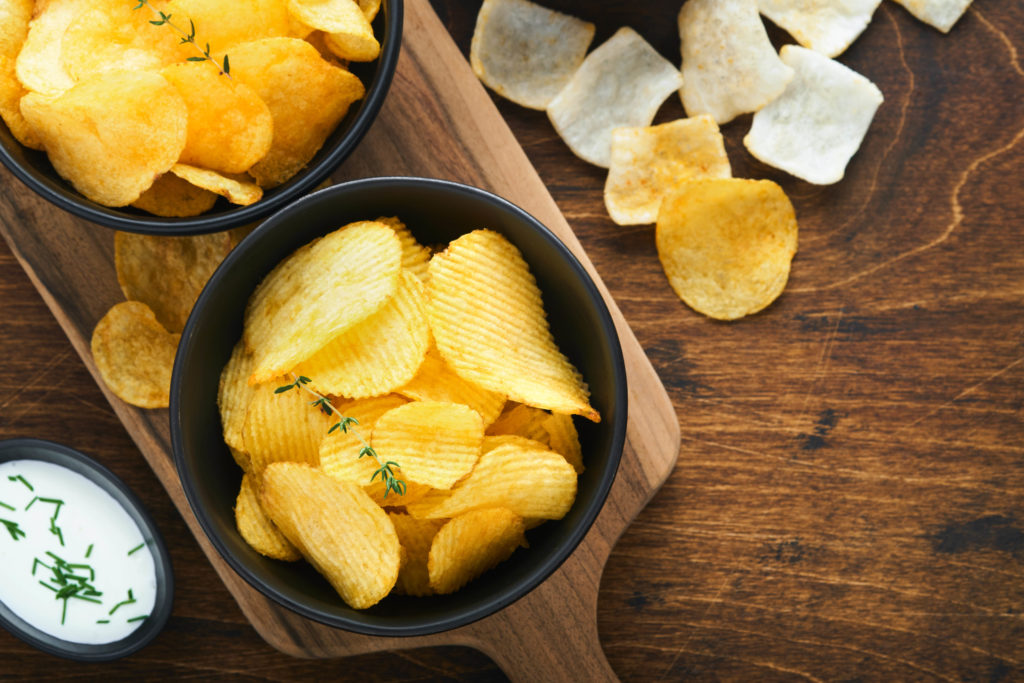 A black bowl holds crispy wavy potato chips with piles of other snack food crisps on a wooden kitchen table similar to the ones in culinary tours with Jaya Travel & Tours
