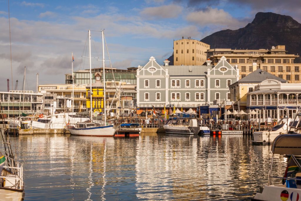 A river filled with boats travel through a town with cute buildings as people walk along the film location for Blood DIamond.