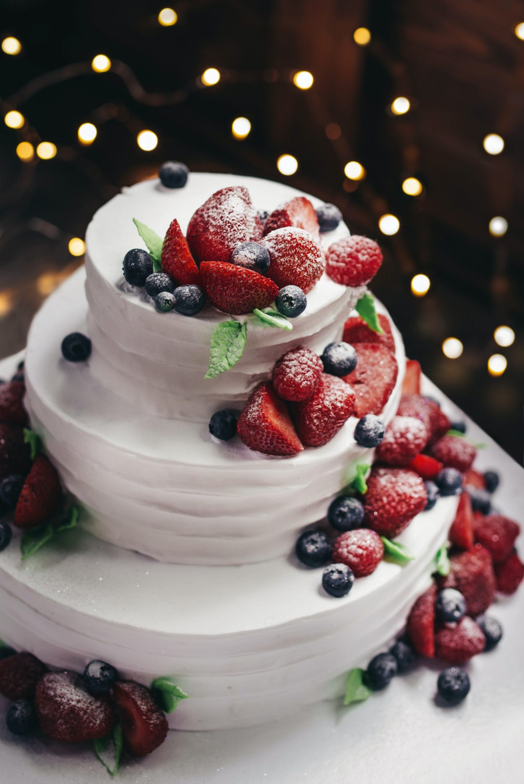 A yummy 3-tier white wedding cake with spatula texture and fresh berries cascading down the side and sprinkled with powdered sugar that you can eat on travel with Jaya.