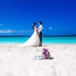A bride and groom in the background with a wedding bouquet in the sand at a Caribbean destination wedding with Jaya Travel & Tours.