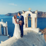 A destination wedding by Jaya Travel & Tours of a young wedding couple on the rooftops of Santorini, Greece with the ocean in the background.