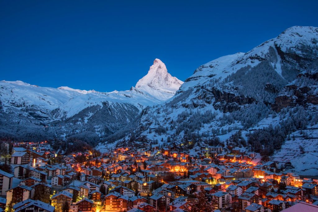 A large mountain covered in snow dominates the picture, right under the blue and starry sky. In the valley is a ski village called Zermatt, Switzerland glows with warm lights all along. A great spring break travel idea from Jaya Travel.