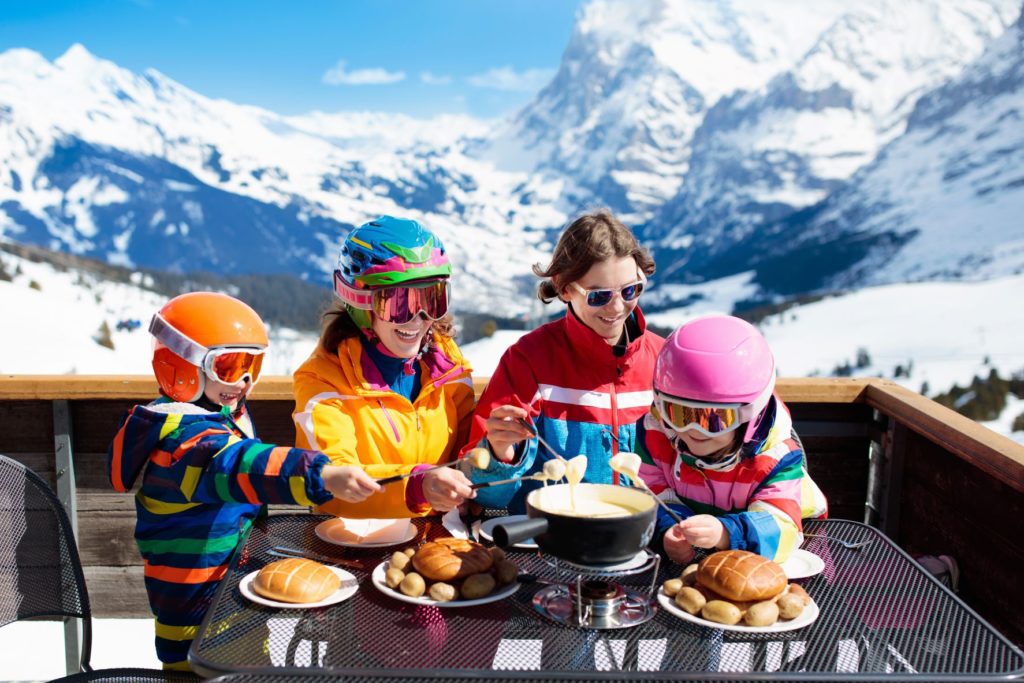 A mother and her three children, all wearing snow clothing and ski protection, eat cheese fondue with bread at a restaurant in Switzerland looking onto the ski slope. Jaya can book this vacation for your spring break!