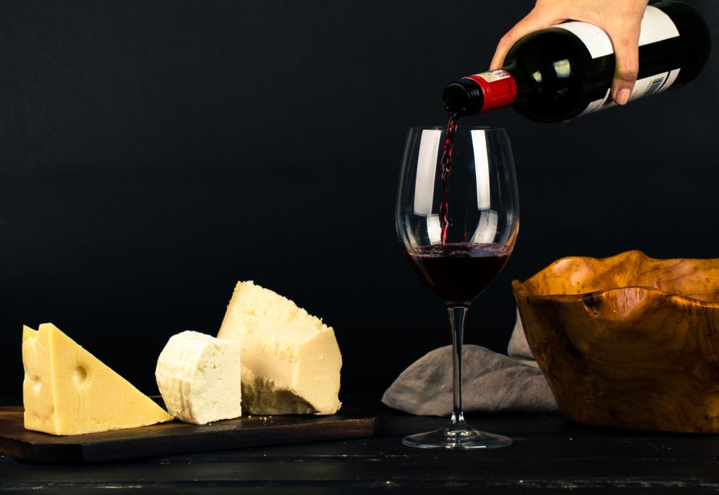 A hand at the top of the frame pours red wine into a tall wine glass sitting on a black table. Next to it is Swiss cheese and bread, which are featured in a culinary tour from Jaya Travel & Tours to Switzerland.
