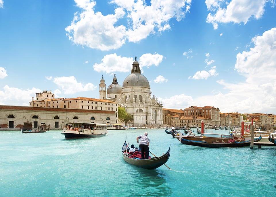 A couple on a romantic vacation ride a gondola boat tour of the Grand Canal with the Basilica Santa Maria in the view. The image is featured in the blog Romantic Valentine's Day Destinations with Jaya Travel & Tours.