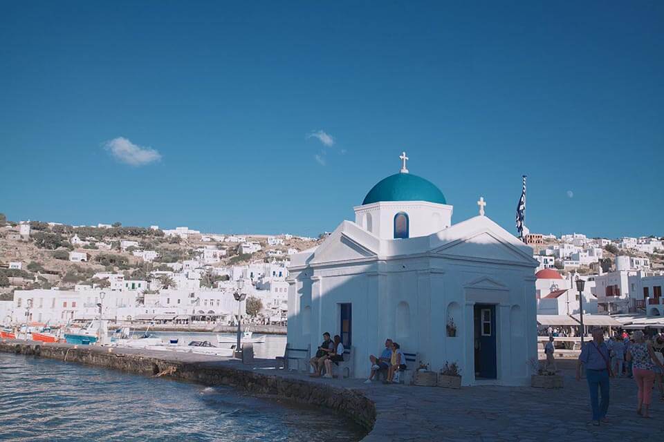 Tourists relax beside one of the iconic blue roof buildings in the romantic neighborhood of Little Venice, on the island of Mykonos, Greece this Valentine's Day Honeymoon destination with Jaya Travel & Tours.