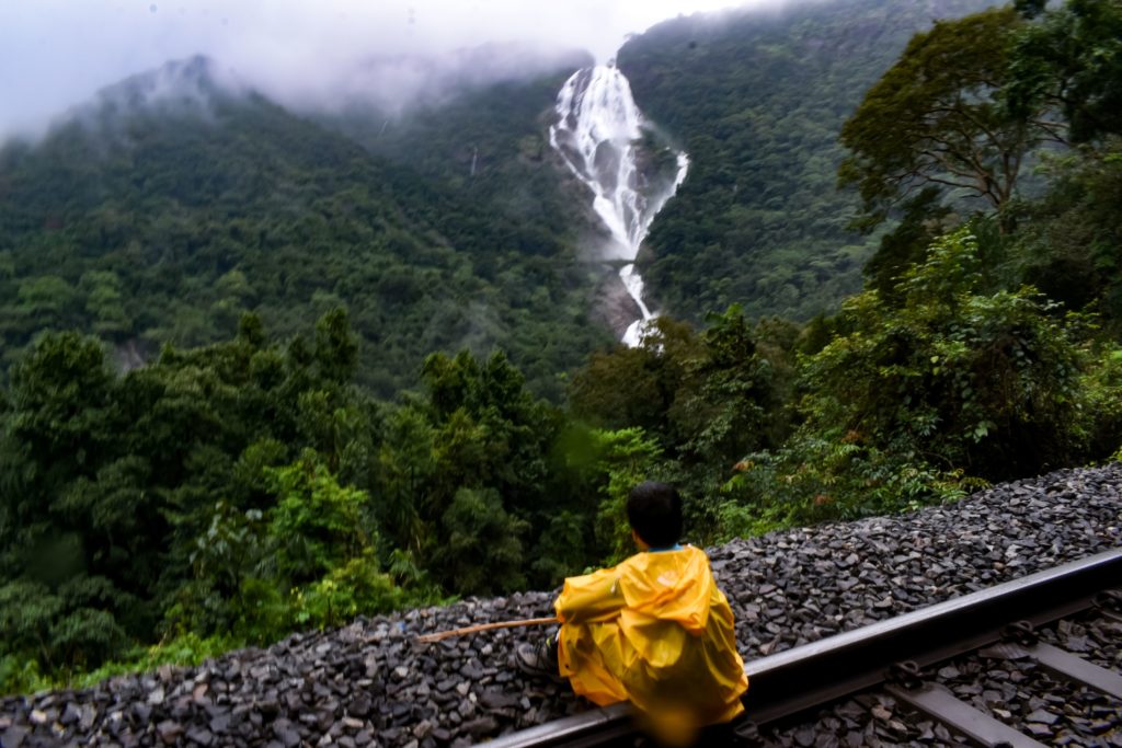 A young boy in a yellow rain coat crouches on an empty railway without trails staring at the beautiful Dudhsagar Waterfalls in India. This image is featured on Jaya Travel & Tours blog article "Which waterfall Should You Visit Quiz?"
