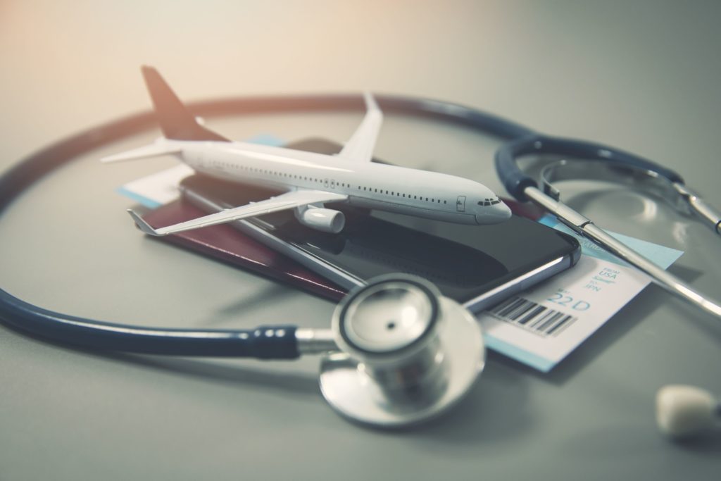 On a white table sits a pile of stacked passports, flight tickets, an airplane, and a stethoscope. The featured image in Jaya Travel & Tours blog, "6 Reasons to Get Travel Insurance," which describes why travelers should buy trip protection.