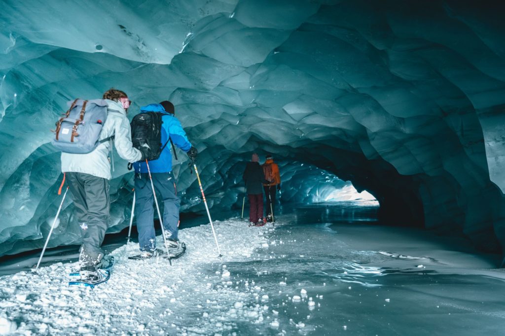 A small tour group of snowshoeing tourists make their way through an iceberg, literally through a tunnel in the center, that is shimmering with beauty. This image is featured in the Jaya Travel & Tours blog, "Which Glacier Should You Visit Quiz," which is a personality travel quiz that determine your next glacier tour.
