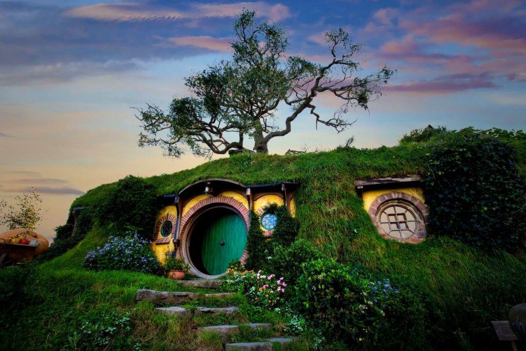 Jaya Travel & Tours travel blog post "On Location: The Lord of the Rings" lists the filming locations from Peter Jackson's popular movie and book trilogy. A tall, grassy hill is the walls of a Hobbit house in Hobbiton in New Zealand, Australia while the sky is darkened with a beautiful sunset.