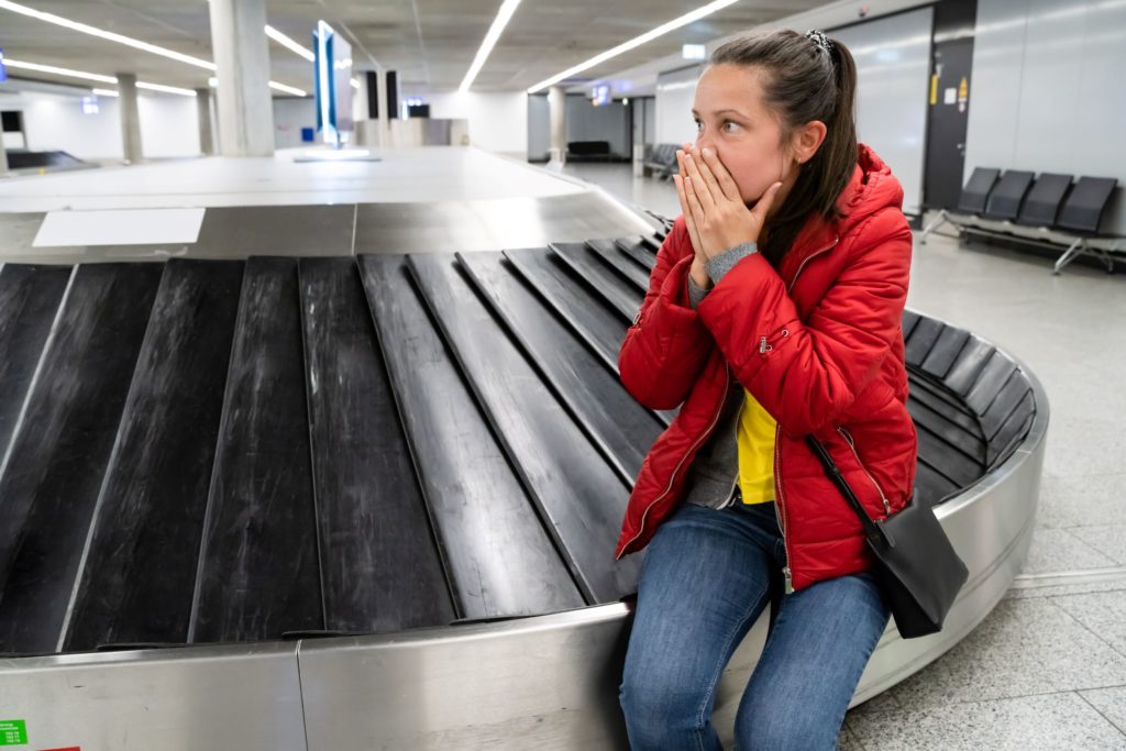 A woman in a red coat sitting on the luggage carousel at the airport is holding her hands to her shocked face because her baggage was lost. In the Jaya Travel article "6 Reasons to Get Travel Insurance," we describe what travel insurance and trip protection can do to protect your vacation and your wallet.