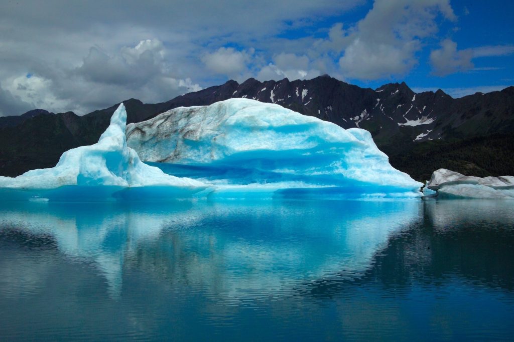 A gigantic bright-blue glacier sits in front of a dark mountain with melted snow and is reflected back in the lake below. This image is featured in the Jaya Travel & Tours blog, "Which Glacier Should You Visit Quiz," which is a personality travel quiz that determine your next glacier tour.