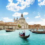 A landscape shot of the Grand Canal and Basilica Santa Maria della Salute in Venice, Italy featured in Jaya Travel & Tours customized tour enchanting italy. The blue water is shining bright from the sun, and several gondolas on the canal are being rowed by men in striped shirts.