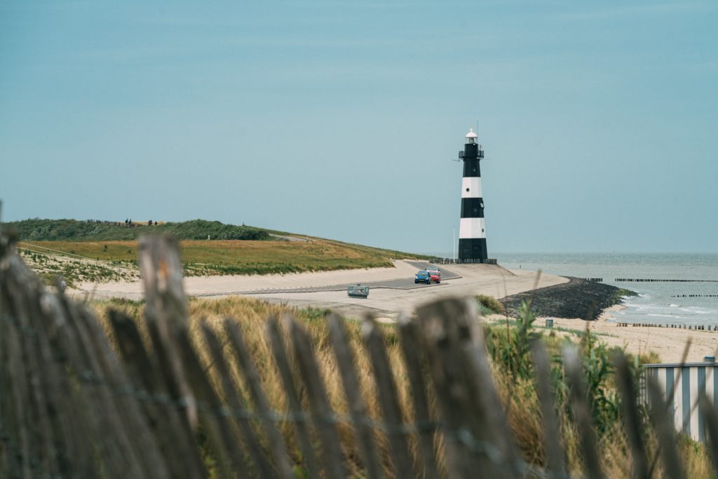 A landscape shot of a black and white lighthouse in Maine with a beach and ocean in the distance, framed by green grass hills and a cloudy sky. This image is featured in the Jaya Travel & Tours blog, "A United States Road Trip," which is a travel guide to the best road trips and driving the best highways in America.