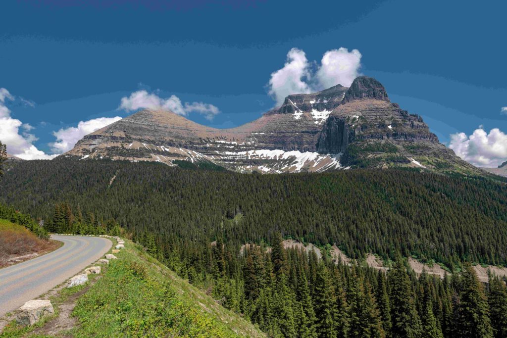 A landscape photograph of Glacier National Park in the USA, with a tall mountain rising over green lush forest, and a small road winding around it. This image is featured in the Jaya Travel & Tours blog, "A United States Road Trip," which is a travel guide to the best road trips and driving the best highways in America.