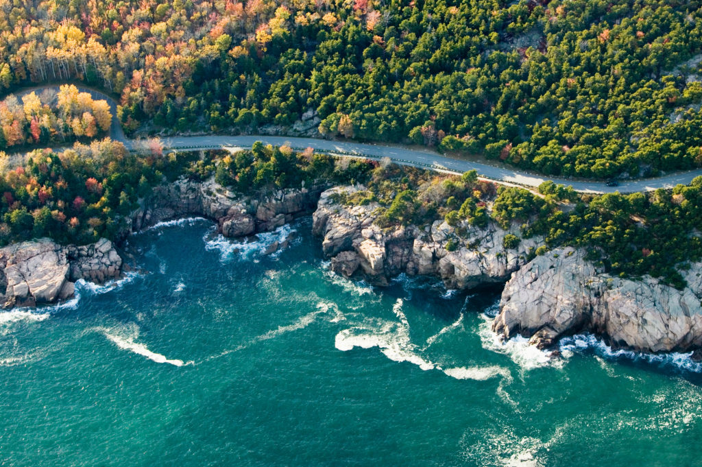 An aerial view of the Maine coastline, forest, and winding road of Arcadia National Park which is being driven on by rental cars. This image is featured in the Jaya Travel & Tours blog, "A United States Road Trip," which is a travel guide to the best road trips and driving the best highways in America.