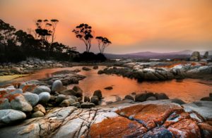 A landscape shot of the Bay of Fires, with a bright orange and yellow sunset overwhelming the sky and reflecting in the water of a bay. This image is featured in the Jaya Travel & Tours blog, "Top 10 Destination: Tasmania," which describes the best destinations to visit in Tasmania and Australia.