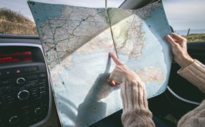 A young woman holds a map away from her chest and points to the coastline where she is taking a road trip with a rental car. This image is featured in the Jaya Travel & Tours blog, "A United States Road Trip," which is a travel guide to the best road trips and driving the best highways in America.