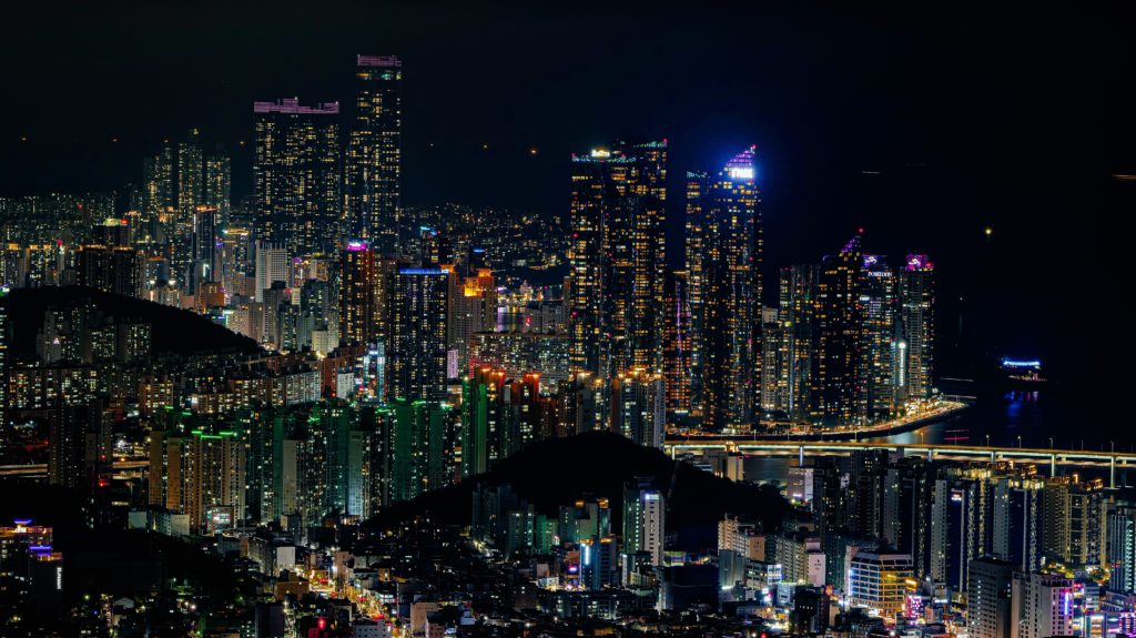 The city of Busan in South Korea at night, which is lit up by bright skyscrapers, apartment lights, road signs, neon lit streets, and more. This image is featured in the Jaya Travel & Tours blog post, "On Location: Black panther & Wakanda Forever," which lists the filming destinations of the popular marvel movie series.