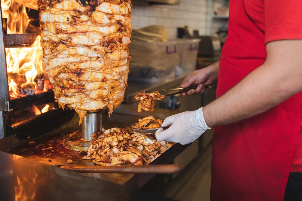 A man stands in front of a large kebob of meat on a spit, which is carved and placed onto a plate with other delicacies. This image is featured in the blog from Sky Bird Travel & Tours, "A Guide To Turkish Cuisine," which describes the types of food, drink, desserts, and dishes from Turkey.