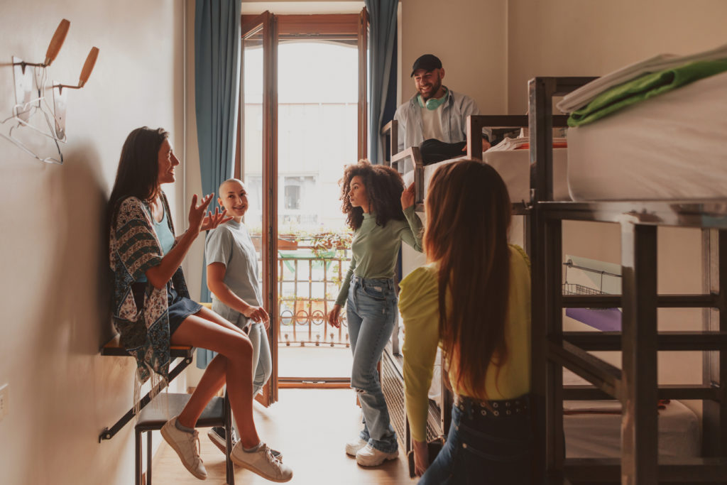 A large group of young people on vacation staying in a hostel together, laughing with each other, and sharing the same room. This image is featured in the Jaya Travel & Tours blog post, "Debunking 5 Myths About Hostels," which describes false lies about staying in a hostel on vacation and why they are incorrect.