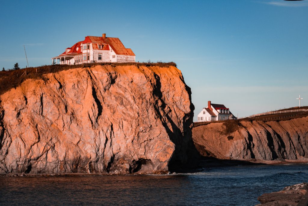 This image of a white and red house built onto a cliff above the ocean is featured in the Jaya Travel & Tours blog, "North America Travel Guide," which describes the best things to do in North America and all of the various destinations.