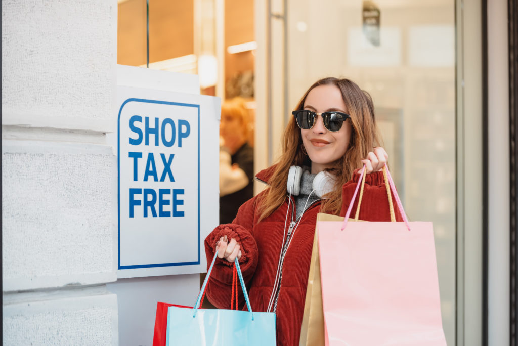 A young woman holding shopping bags walking by a sign that says "shop tax free" is featured in the Jaya Travel & Tours travel guide blog post, "How to Travel Tax-Free Internationally," which describes the methods that people can take to save money on vacation.