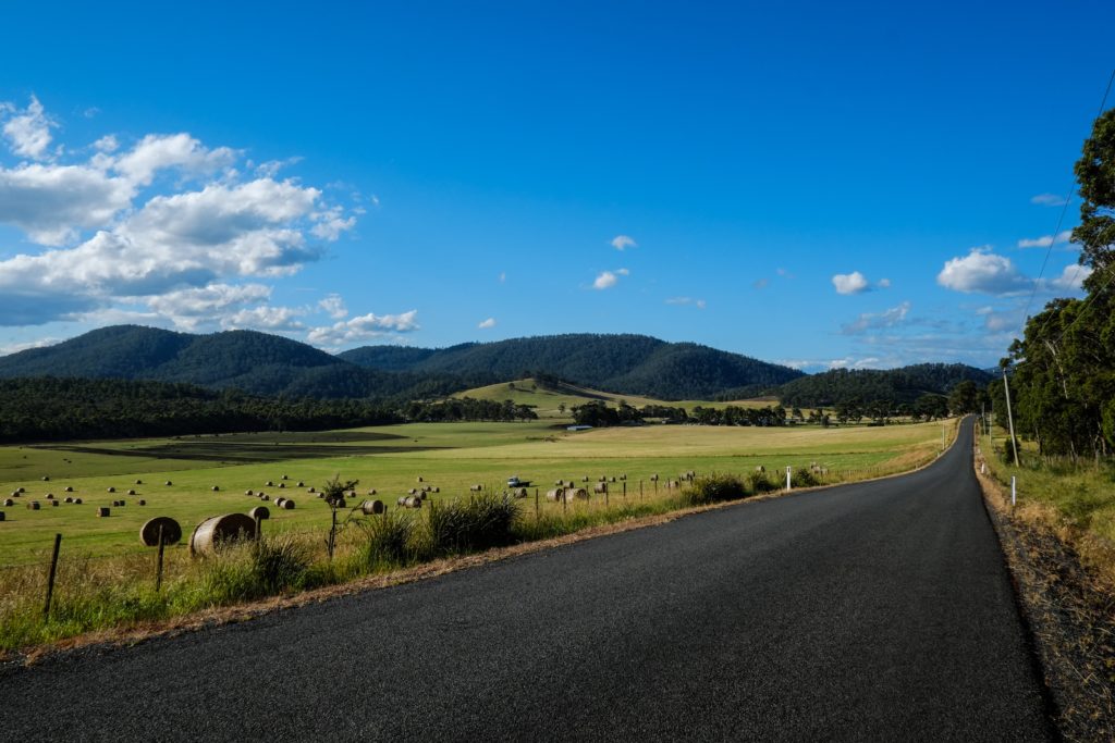 This image of a road running through a mountain range next to a field of hay is featured in the Jaya Travel & Tours blog post "Australia Travel Guide"
