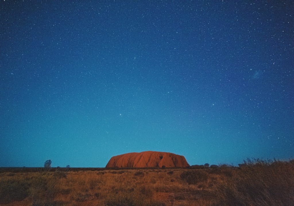 This image of a rock formation in the desert at night with bright stars in the sky is featured in the Jaya Travel & Tours blog post "Australia Travel Guide"