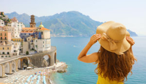 Featured in Best Destinations for Solo Vacations by Jaya Travel & Tours, this image shows Summer holiday in Italy with the back view of young woman holding her hat with Atrani village on the background, Amalfi Coast, Italy