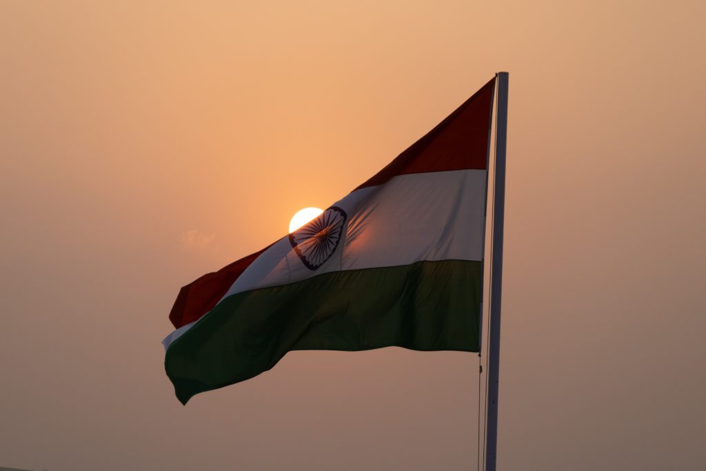 Featured in India Travel Guide by Jaya Travel & Tours, this image shows the Indian flag waving with a beautiful sunset behind.