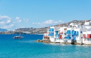 Mykonos - Jaya Travel & Tours Whitewashed buildings with blue balconies line the rocky shoreline, turquoise water lapping the coast, a sailboat in the distance under a clear sky, daytime.