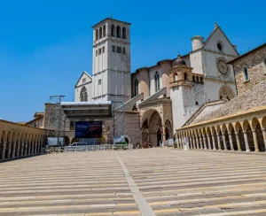 basilica-di-san-francesco-in-assisi - Jaya Travel & Tours Sunlit historic cathedral with a tall bell tower and arched cloister surrounding a wide stone plaza with a temporary stage nearby.