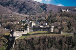 castelgrande-castle-bellinzona - Jaya Travel & Tours Medieval stone castle with crenellated walls and towers on a hillside overlooking a small village and forested valley.
