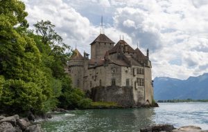chillon-castle - Jaya Travel & Tours Medieval stone castle on a rocky lakeshore with trees to the left and distant mountains in the background on a calm lake.