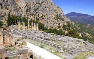 delphi - Jaya Travel & Tours Ancient stone ruins with tall columns on a terraced plaza, set on a hillside with mountains in the background and clear blue sky.