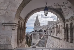 fisherman-bastion - Jaya Travel & Tours Stone arch frames a medieval fortress terrace with a tall conical tower, staircases, and visitors overlooking the city.