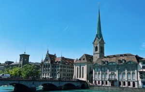 fraumunster-church - Jaya Travel & Tours Historic European cityscape along a river with a stone arched bridge and a tall church spire.