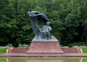 fryderyk-chopin-monument - Jaya Travel & Tours Bronze statue of a seated figure beneath a flowing draped arch, on a red stone pedestal in a tree-filled park with a reflecting pool nearby in front.