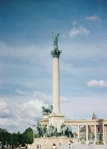 heroes-square - Jaya Travel & Tours Tall monument: a high column crowned by a statue with wings, surrounded by bronze figures and horses in a plaza setting.