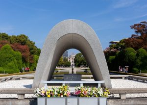 hiroshima-peace-memorial-museum - Jaya Travel & Tours Front view of a large gray concrete arch memorial with a hollow center, in a landscaped park with flowers at its base.