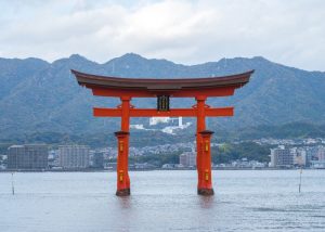 itsukushima-shrine - Jaya Travel & Tours Red torii gate standing in calm water with a city and forested mountains in the background, under a cloudy sky.