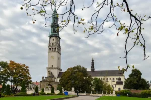 jasna-gora-monastery - Jaya Travel & Tours Historic church with a tall clock tower and green copper spire, framed by autumn trees in a park-like setting.
