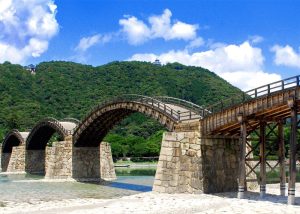 kintaikyo-bridge - Jaya Travel & Tours Traditional arched wooden bridge over a shallow river, supported by stone piers, with green hills and a blue sky in the background.